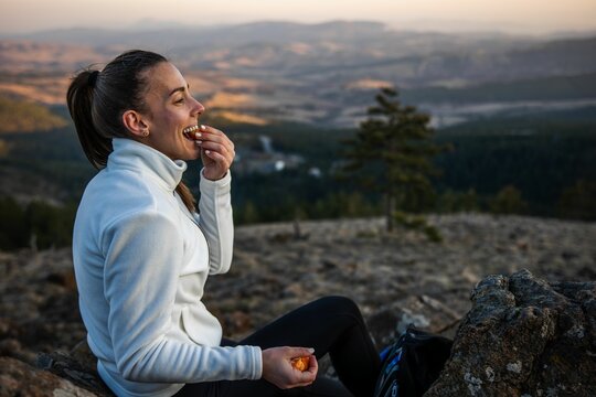 Woman in a white jacket enjoys a snack while sitting on a mountain with a scenic view at sunset.