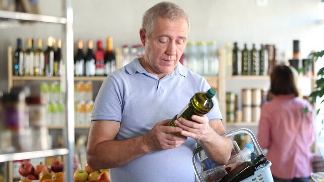 Mature man buyer looks thoughtfully at bottles of olive oil, woman client in background. He choose olive oil. Client examines product in detail, get acquainted with information for consumer