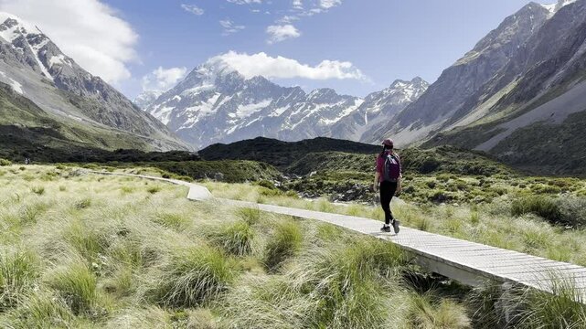 backpacker girl hikes on wooden boardwalk overlooking famous tall mount cook (aoraki) on hooker valley track in Mount cook national park south island New Zealand. ost popular trail on south island