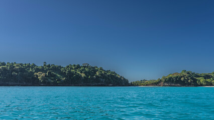 Fototapeta premium The coast of a tropical island. View from the ocean. Ripples on the calm aquamarine surface of the sea water. Silhouettes of people on a sandy beach. Green hills against a clear blue sky. Madagascar.