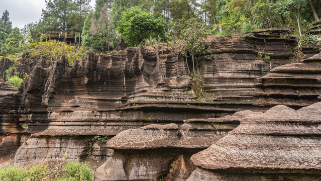 Amazing red-brown karst rocks. Steep layered slopes with a wavy surface. Green vegetation on the cliffs and in the valley. An observation deck in the distance. China. Red Stone Forest National Geopark