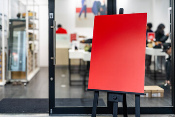 Red display board standing on easel outside shop entrance with blurred people and modern interior...