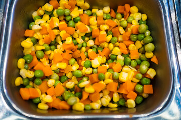 Close-up of mixed steamed vegetables including corn, carrots, and peas in a stainless steel serving tray