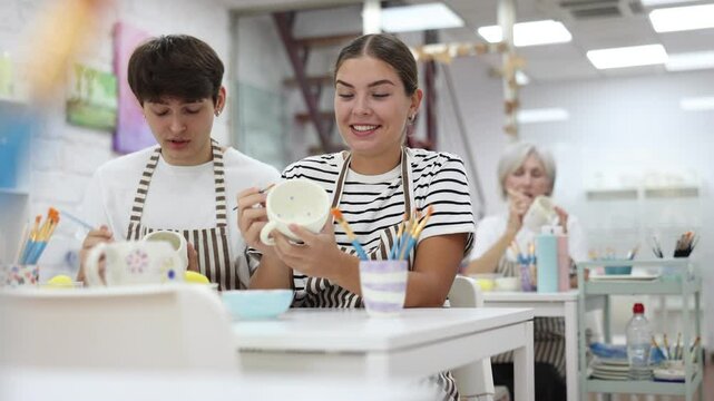 Cheerful young couple, guy and girl, actively engaged in creative hobby, decorating ceramic pottery together at table in cozy workshop setting