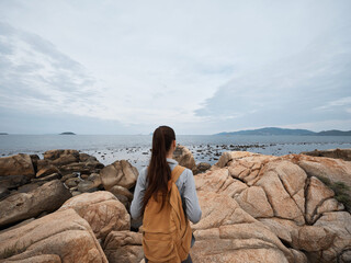 Young woman exploring rocky coast during overcast day, wearing casual clothes and backpack,...
