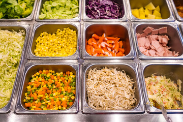 Top view of a salad bar with various fresh ingredients including vegetables, tuna, corn, carrots, purple cabbage, and pineapple