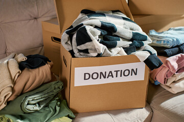 Open donation box filled with donated clothes in living room. Cardboard box with used clothes for...