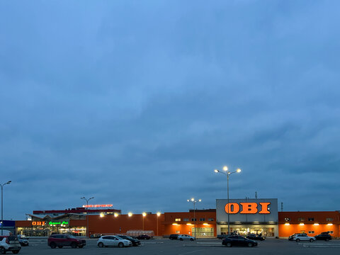 Exterior view of a large OBI DIY hypermarket building with an illuminated bright orange brand logo at dusk.