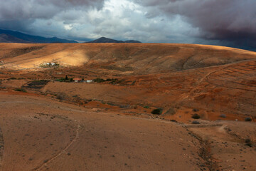 Dramatic storm clouds gather over Fuerteventura's arid volcanic landscape with golden hills and distant mountains. Small rural settlement nestled in valley with dirt roads crossing the characteristic 