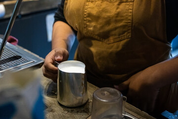Asian barista in brown apron holds metal pitcher of steamed milk beside espresso machine in cozy café, preparing drink with focus, showing hands-on coffee-making process