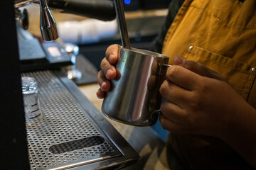 Asian barista wearing brown apron holds metal pitcher under steam wand to froth milk beside espresso machine in cozy café, showcasing hands-on coffee preparation and barista technique