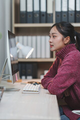Young woman in a cozy sweater concentrating on a computer screen at a modern office desk. Shelves...