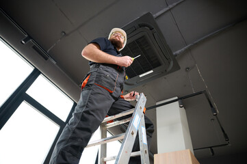 Bearded man staying on ladder installing air conditioner at the office. Ceiling mounted cassette air conditioner in modern building. Loft style. Industrial interior design. Technician servicing © Василь Івасюк