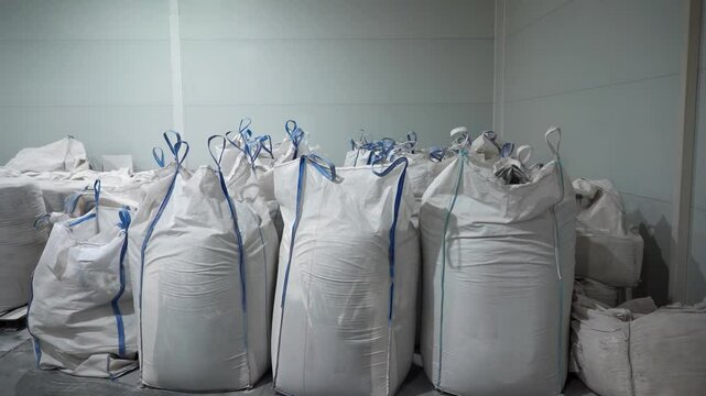 Big bags with microplastic granules stored in empty warehouse. Recyclable polyester materials in large sacks on manufacturing plant storage floor