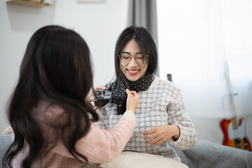 Fototapeta premium Smiling woman receiving scarf from friend indoors, cozy home setting, intimate moment showcasing fashion and comfort with warm colors and soft light