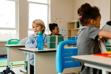 Obraz premium A schoolboy sits at a desk and looks away while a classmate sits in front of him and a teacher sits behind him, at school, in the classroom