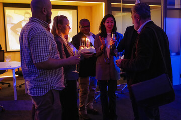 Group of seven employees standing and holding glasses, cake with candles, and bottle, during retirement celebration, at work