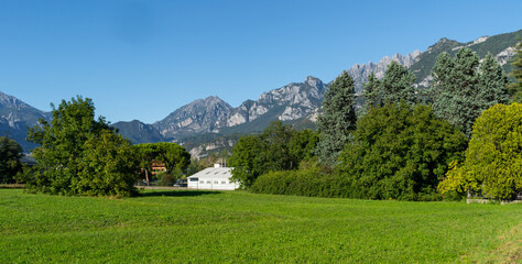 Vibrant green field surrounded by trees and distant mountains near Lecco, lake Como, Italy. Small building peeks through the foliage, rural scene with natural beauty of mountainous landscape