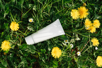 White cosmetic tube lies in green grass surrounded by yellow dandelions, cream product packaging, sunscreen tube mock up, eco friendly concept, white bottle creme