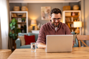 Happy man working remotely on his laptop in a cozy home office. Warm lighting, bookshelves, and...