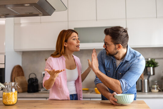 Intense disagreement between a couple in their kitchen. The man raises his hand in a defensive manner. Both display strong emotions, reflecting a challenging moment in their relationship - Powered by Adobe