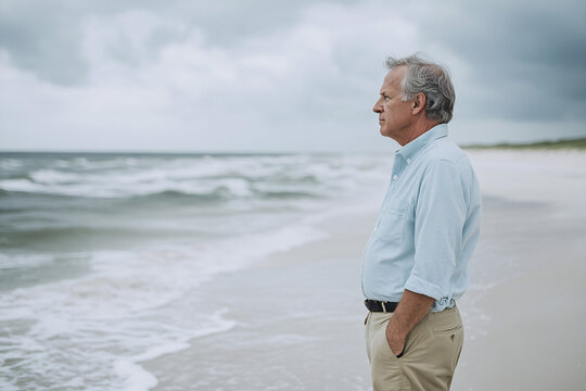A mature man in a light blue shirt and khaki pants stands on the beach hands in pockets gazing at the ocean with dark clouds in the sky
