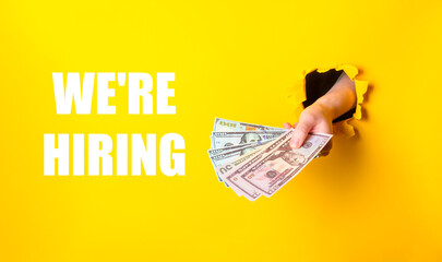 Hand holding a stack of money with the words We're hiring written below it. The image has a bright...