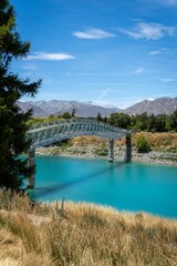Obraz premium A scenic view of a turquoise river with a metal bridge and mountains at Tekapo, New Zealand.