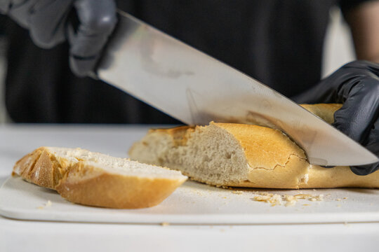 chef wearing black gloves slicing fresh baguette with a sharp knife on a white cutting board, with crispy golden crust and soft fluffy texture inside