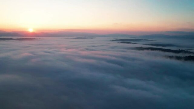 Aerial view of sun rises oversea of clouds, casting gentle pink hue across sky. Mountain peaks and tree tops poke through mist, creating ethereal, dream-like landscape of rolling fog, distant horizons