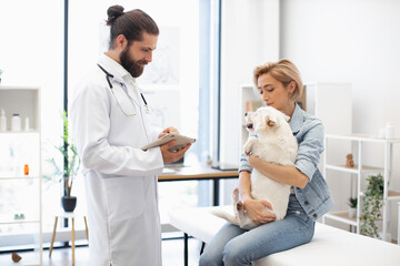 Male veterinarian in white coat examines dog held by young adult woman at clinic. Modern light...