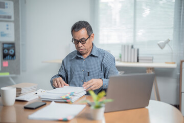 Fototapeta premium Focused mature employee working with documents and using laptop, managing paperwork and preparing financial report while sitting at desk in modern office