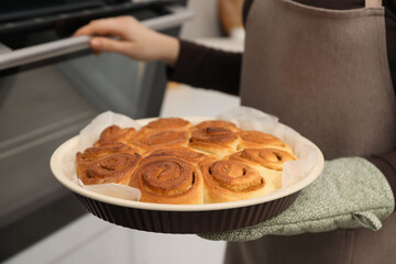Woman with delicious cinnamon rolls near oven indoors, closeup