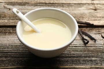 Tasty condensed milk, spoon and vanilla pods on wooden table, closeup