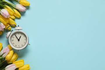 Spring time. Alarm clock and beautiful tulips on light blue background, flat lay. Space for text