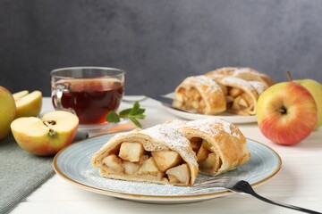 Pieces of tasty apple strudel with powdered sugar and fork on white wooden table against grey background, closeup