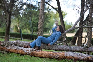 Young, beautiful, brunette, Hispanic woman, wearing a denim suit, relaxed and calm looking at the camera, lying on the fallen trunk of a tree, in nature. Concept fashion, beauty, trendy, femininity.