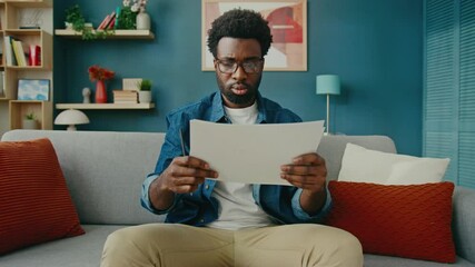 African American man in glasses carefully examining two documents with graphs. Analyzing financial data or business reports in modern home office. Remote work or strategy planning concept.