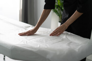 In a contemporary beauty salon, a cosmetologist arranges a clean treatment couch with fresh linens,...