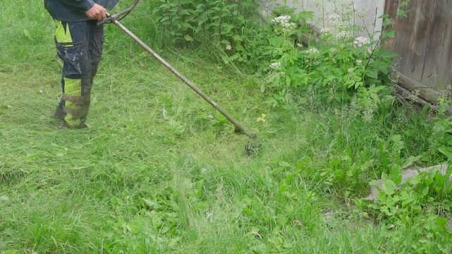 Gardener in protective gear using a string trimmer to cut tall grass and weeds near a building.
