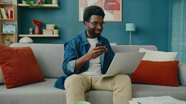 Positive African American man wearing glasses sitting on modern sofa using laptop. Holding credit card while completing online transaction. Feeling satisfied while shopping or managing finances.