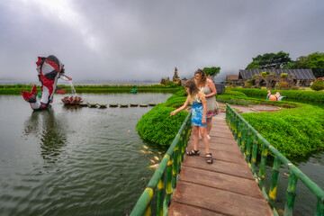 A joyful family strolls happily across a beautiful wooden bridge spanning a tranquil pond, embraced by lush greenery and delightful sculptures in Ulun Danu Beratan Temple, Bali, Indonesia