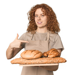 Young Caucasian redhead woman holding bread tray person pointing by hand to a shirt copy space,...