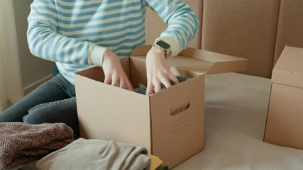 Woman packs donation box with clothes at home. Person places warm sweater into cardboard box. Preparing used clothing for recycling or second hand. Concept of charity and humanitarian aid