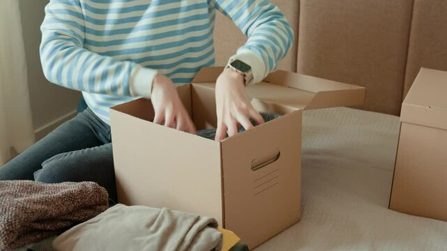 Woman packs donation box with clothes at home. Person places warm sweater into cardboard box. Preparing used clothing for recycling or second hand. Concept of charity and humanitarian aid