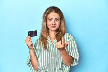 Fototapeta premium Young blonde with a credit card in blue studio pointing with finger at you as if inviting come closer.