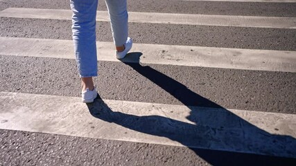 Female legs crossing pedestrian crossing, wearing blue jeans and white sneakers during bright summer day, showcasing urban street walking with safety and motion