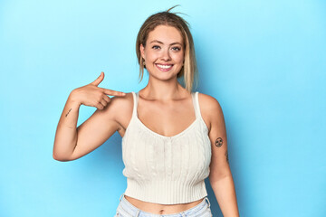 Blonde young woman in white top on blue background person pointing by hand to a shirt copy space,...