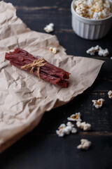Dry red beef, jerky meat sticks tied with jute thread on paper and black wooden background. Bowl of popcorn. Copy space, vertical shot
