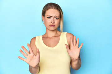 Fototapeta premium Blonde young woman in yellow tank top on blue background rejecting someone showing a gesture of disgust.